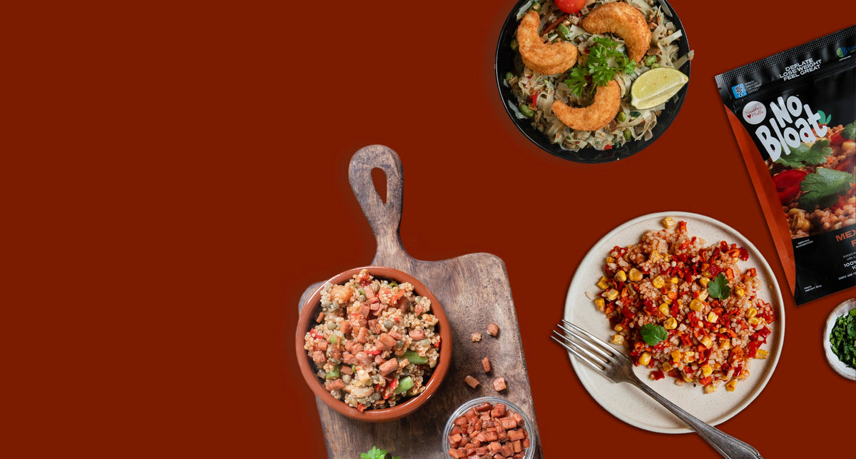 A styled food presentation on a solid dark red background, featuring a bowl of vegan prawns and mushroom edamame, a plate of mexican rice, a bowl of vegan bacon cubes and 'No Bloat' packet on the right.