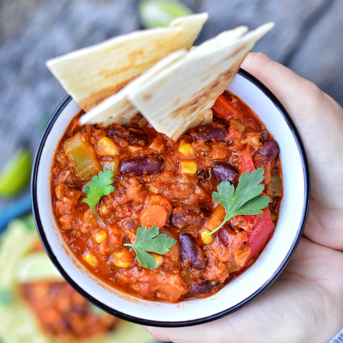 A close-up of a hand holding a white bowl with a black rim filled with a chunky chilli bean dip and tortilla chips resting on the rim.