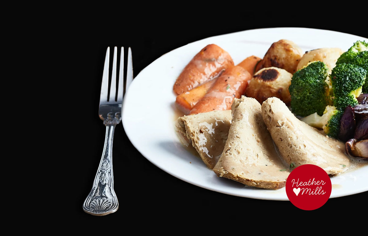A white plate of vegan turkey roast on a black background, including, carrots, green broccoli, purple onions, and roasted potatoes, with a silver fork to the left.