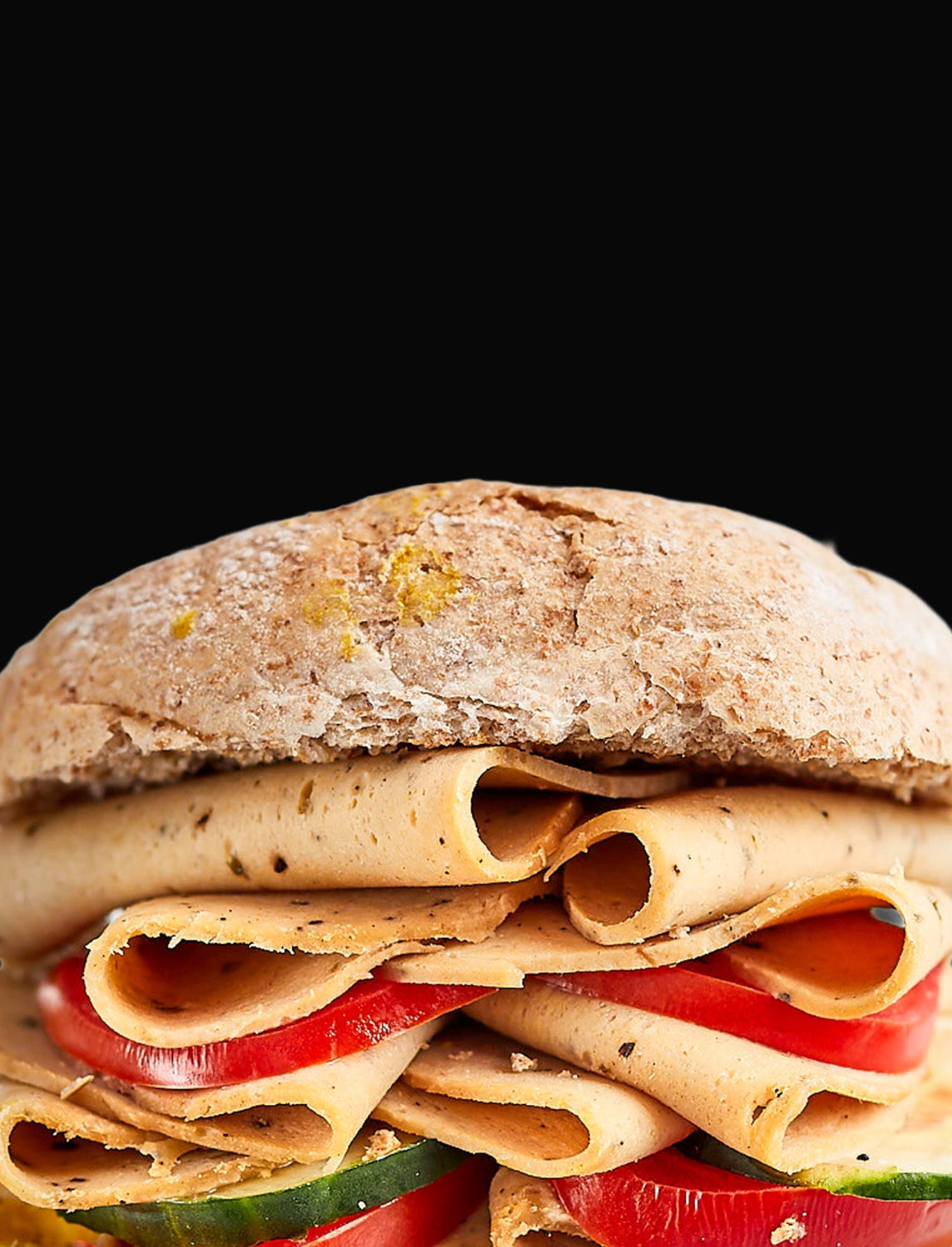 A close-up of a sandwich with a rustic whole wheat bread bun, layered with sliced sage and onion, red bell peppers, and cucumber, set against a solid black background.