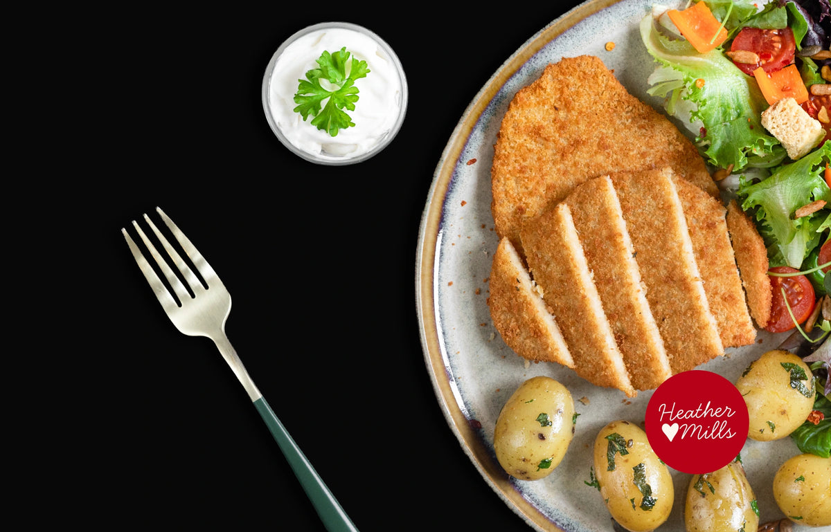 A plated meal featuring two thick, golden-brown breaded schnitzels served with small boiled potatoes, a side salad with tomatoes and carrots, and a small bowl of white sauce on a black background. A red circular logo overlay reads 'Heather Mills'.